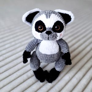 May include: A gray and white crocheted lemur plush toy with black eyes and black ears. The lemur is sitting on a white surface.