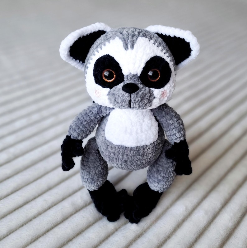 May include: A gray and white crocheted lemur plush toy with black eyes and black ears. The lemur is sitting on a white surface.