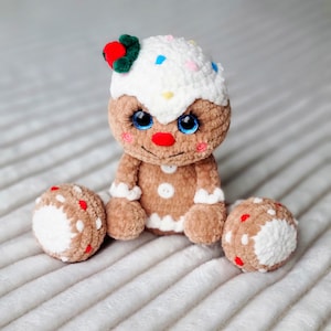 May include: A crocheted gingerbread man toy with white icing, red and white sprinkles, and blue eyes. The toy is sitting on a white fluffy surface.