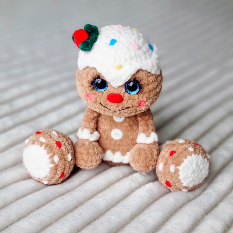 May include: A crocheted gingerbread man toy with white icing, red and white sprinkles, and blue eyes. The toy is sitting on a white fluffy surface.