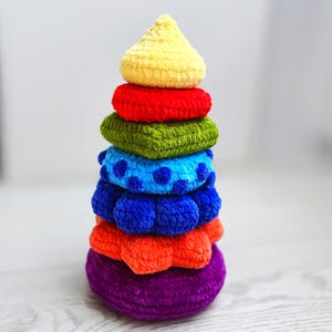 Puede incluir: Un juguete apilable de ganchillo hecho a mano, colorido. El juguete presenta una base morada, seguida de naranja, azul, azul con lunares, verde, rojo y una parte superior amarilla. Un juguete suave y afelpado para bebés y niños pequeños.