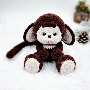 May include: A brown crocheted monkey plush toy with white accents on the face and feet. The monkey is sitting on a white fluffy surface.