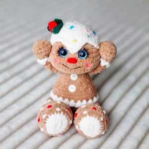 May include: A crocheted gingerbread person toy with white icing, red and green candy decorations, and blue eyes. The toy is sitting on a white and brown striped surface.