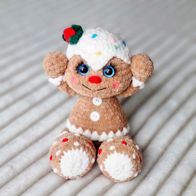 May include: A crocheted gingerbread person toy with white icing, red and green candy decorations, and blue eyes. The toy is sitting on a white and brown striped surface.