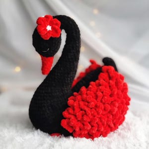 Puede incluir: Una figura de cisne negro hecha a mano con una flor roja en la cabeza y detalles rojos en las alas. El cisne está hecho de un material suave y texturizado y se encuentra sobre una superficie blanca. Un artículo decorativo.