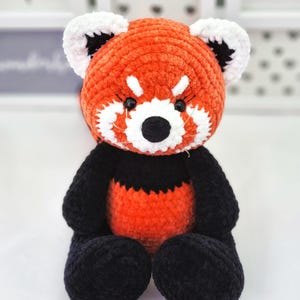 May include: A crocheted red panda plush toy with black and white facial features. The toy is sitting on a white surface.