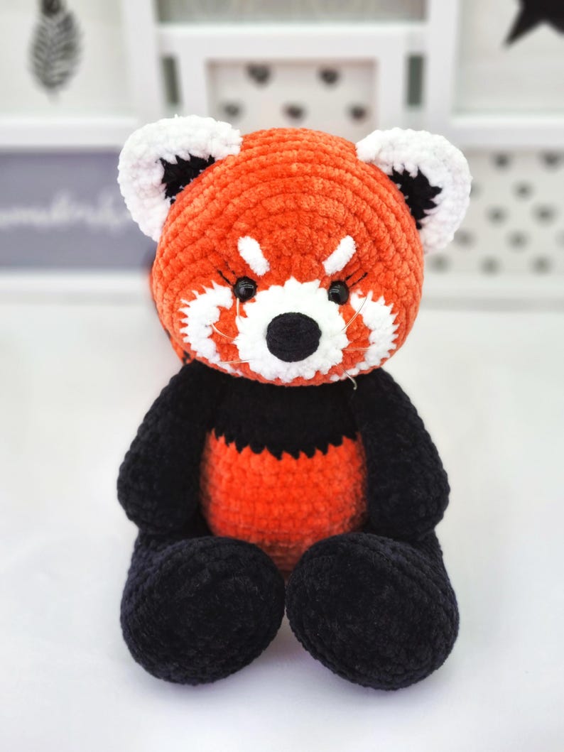 May include: A crocheted red panda plush toy with black and white facial features. The toy is sitting on a white surface.