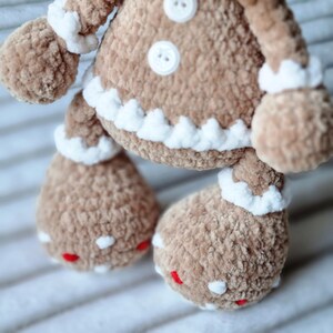 May include: A brown crocheted gingerbread man toy with white icing details and red buttons. The toy has a white crocheted trim around the edges of the body and arms.