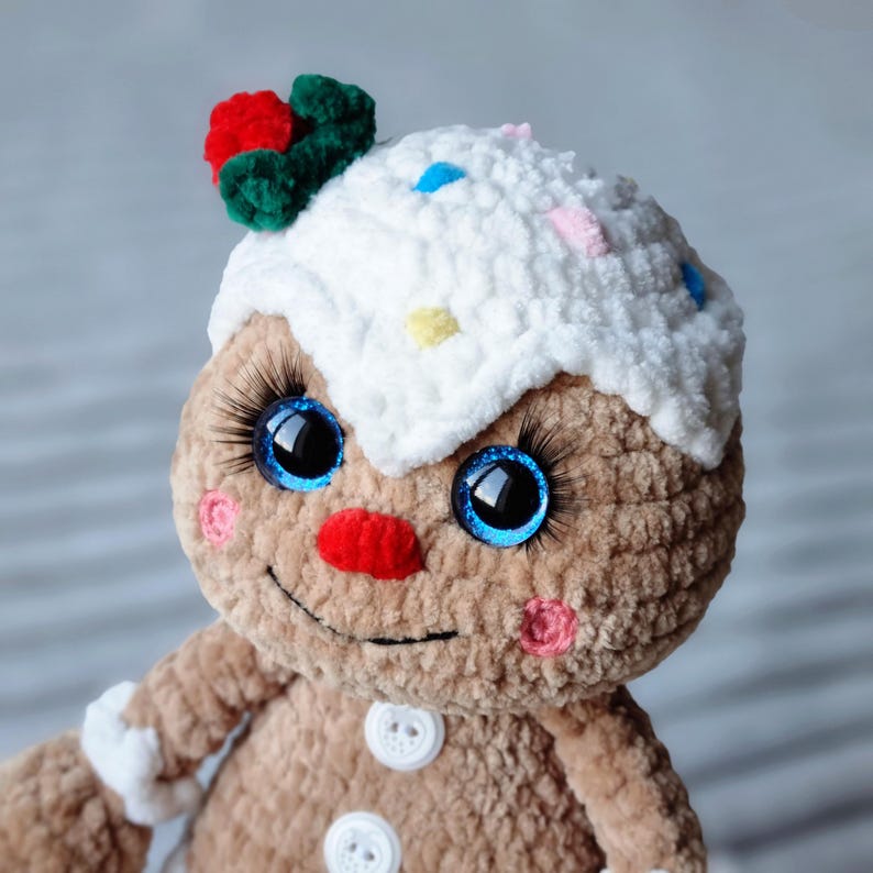 May include: A crocheted gingerbread man toy with a white icing hat, blue eyes, and a red nose. The hat is decorated with colorful sprinkles.