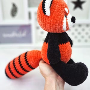May include: A crocheted red panda plush toy with black and orange striped tail. The toy is made with soft, fuzzy yarn and has a white face with black eyes and a black nose.