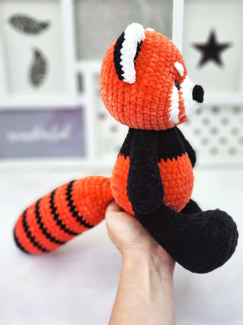 May include: A crocheted red panda plush toy with black and orange striped tail. The toy is made with soft, fuzzy yarn and has a white face with black eyes and a black nose.