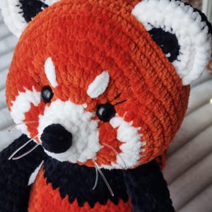 May include: A crocheted red panda plush toy with black and white details. The toy has black eyes, a black nose, and white fur around the eyes and mouth.