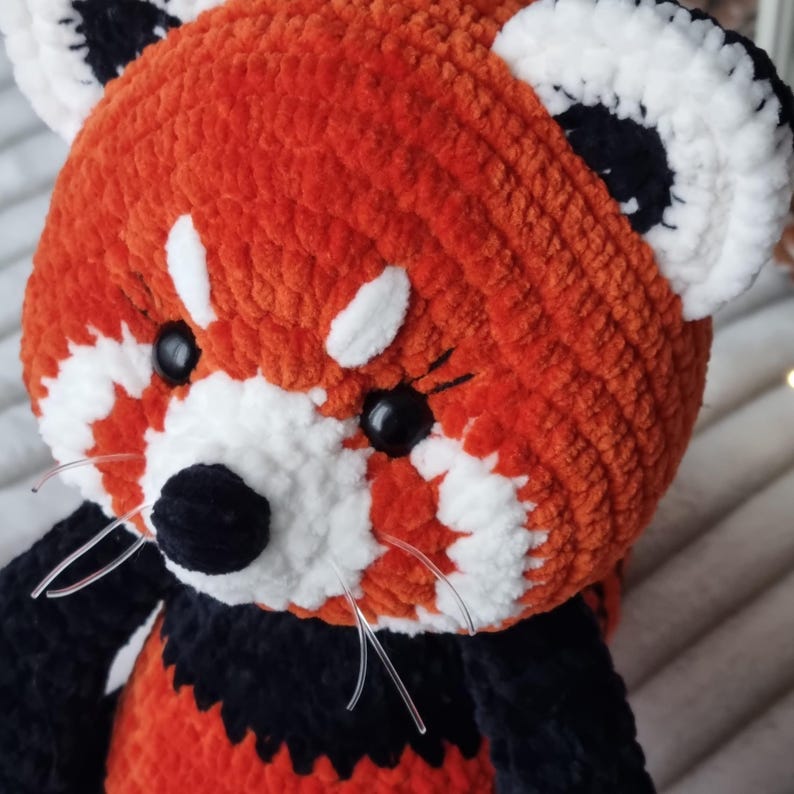 May include: A crocheted red panda plush toy with black and white details. The toy has black eyes, a black nose, and white fur around the eyes and mouth.