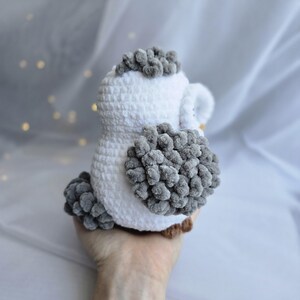 Pu&ograve; includere: Un gufo all'uncinetto fatto a mano, bianco e grigio. Il gufo ha una consistenza soffice con accenti grigi sulla testa, sulle ali e sulla coda. Il gufo &egrave; tenuto in mano, mostrando il design morbido e peluche. I piedi del gufo sono marroni.