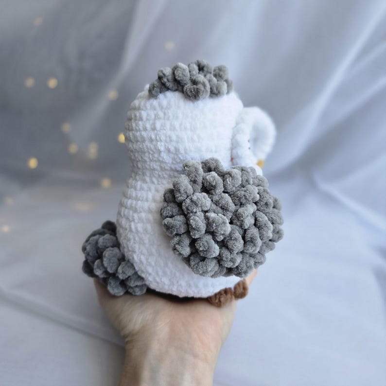 Pu&ograve; includere: Un gufo all'uncinetto fatto a mano, bianco e grigio. Il gufo ha una consistenza soffice con accenti grigi sulla testa, sulle ali e sulla coda. Il gufo &egrave; tenuto in mano, mostrando il design morbido e peluche. I piedi del gufo sono marroni.