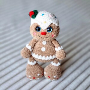 May include: A gingerbread man plush toy with white icing, red and green decorations, and blue eyes. The toy is standing on a white and gray striped surface.