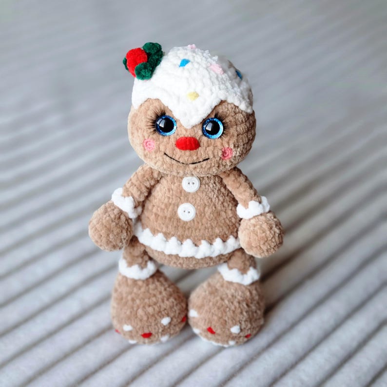 May include: A gingerbread man plush toy with white icing, red and green decorations, and blue eyes. The toy is standing on a white and gray striped surface.