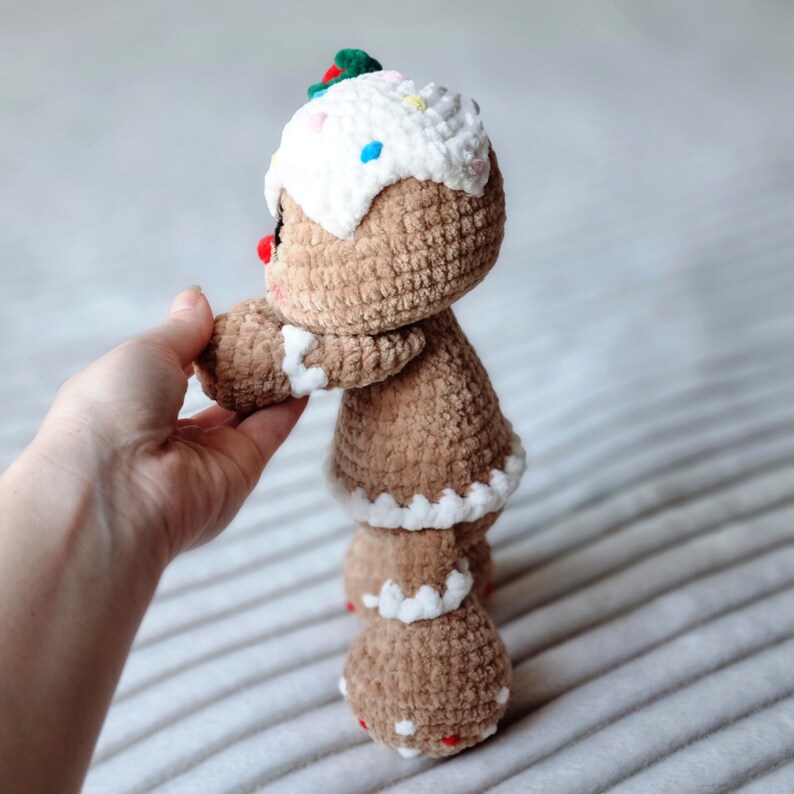 May include: A crocheted gingerbread person toy with brown fur, white icing, and colorful sprinkles. The toy is standing on a white and brown striped surface.