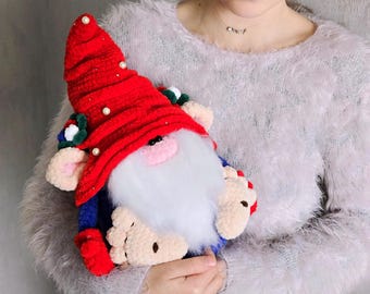 May include: A handmade gnome doll with a red hat adorned with white pearls and a white beard. The gnome has a blue body, beige feet, and is held by a person wearing a fluffy gray sweater. The doll is a festive decoration.