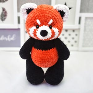 May include: A crocheted red panda plush toy with black and white details. The toy has a red body, black legs and arms, and a white face with black eyes and a black nose.