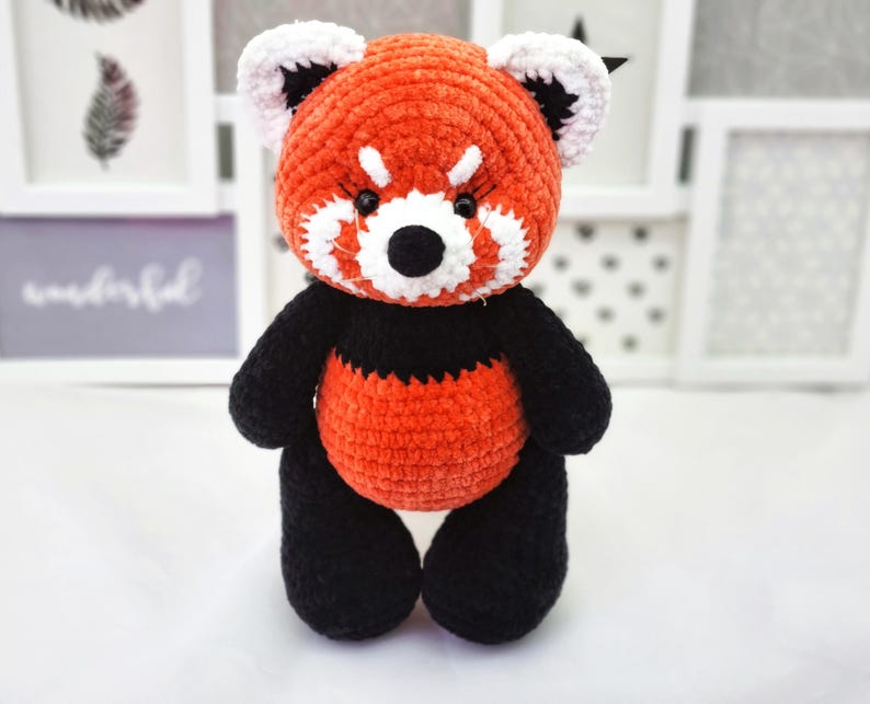 May include: A crocheted red panda plush toy with black and white details. The toy has a red body, black legs and arms, and a white face with black eyes and a black nose.