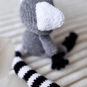 May include: A crocheted gray and white lemur toy with a black and white striped tail. The lemur has large white ears and a small black nose.