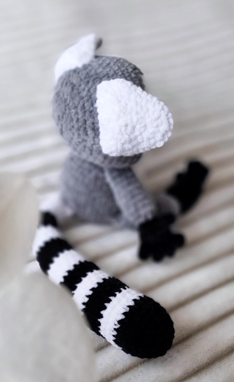 May include: A crocheted gray and white lemur toy with a black and white striped tail. The lemur has large white ears and a small black nose.