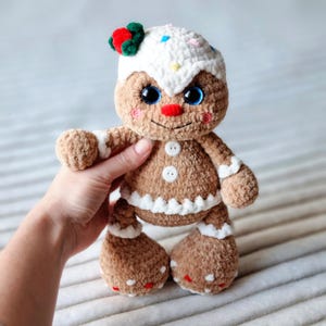 May include: A crocheted gingerbread man toy with a white icing hat, red and green candy decorations, and button eyes.