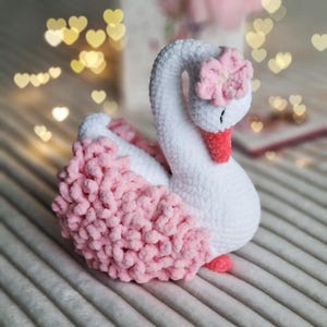 Puede incluir: Un cisne blanco de crochet con una flor rosa en la cabeza. El cisne tiene una cola rosa y esponjosa.