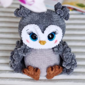 Op de afbeelding: Een handgemaakte gehaakte uilpop in grijs en wit met grote blauwe ogen, lange wimpers en een kleine gele snavel. De uil heeft bruine poten en kleine plukjes garen op zijn hoofd, waardoor hij een pluizig uiterlijk heeft.