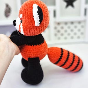 May include: A crocheted red panda plush toy with black and white details. The toy has a long, striped tail and is standing on a white surface.