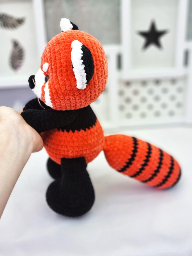 May include: A crocheted red panda plush toy with black and white details. The toy has a long, striped tail and is standing on a white surface.