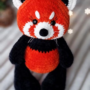 May include: A crocheted red panda plush toy with black and white details. The toy has a red body, black legs and ears, and white facial markings. The toy is sitting on a white fuzzy surface.
