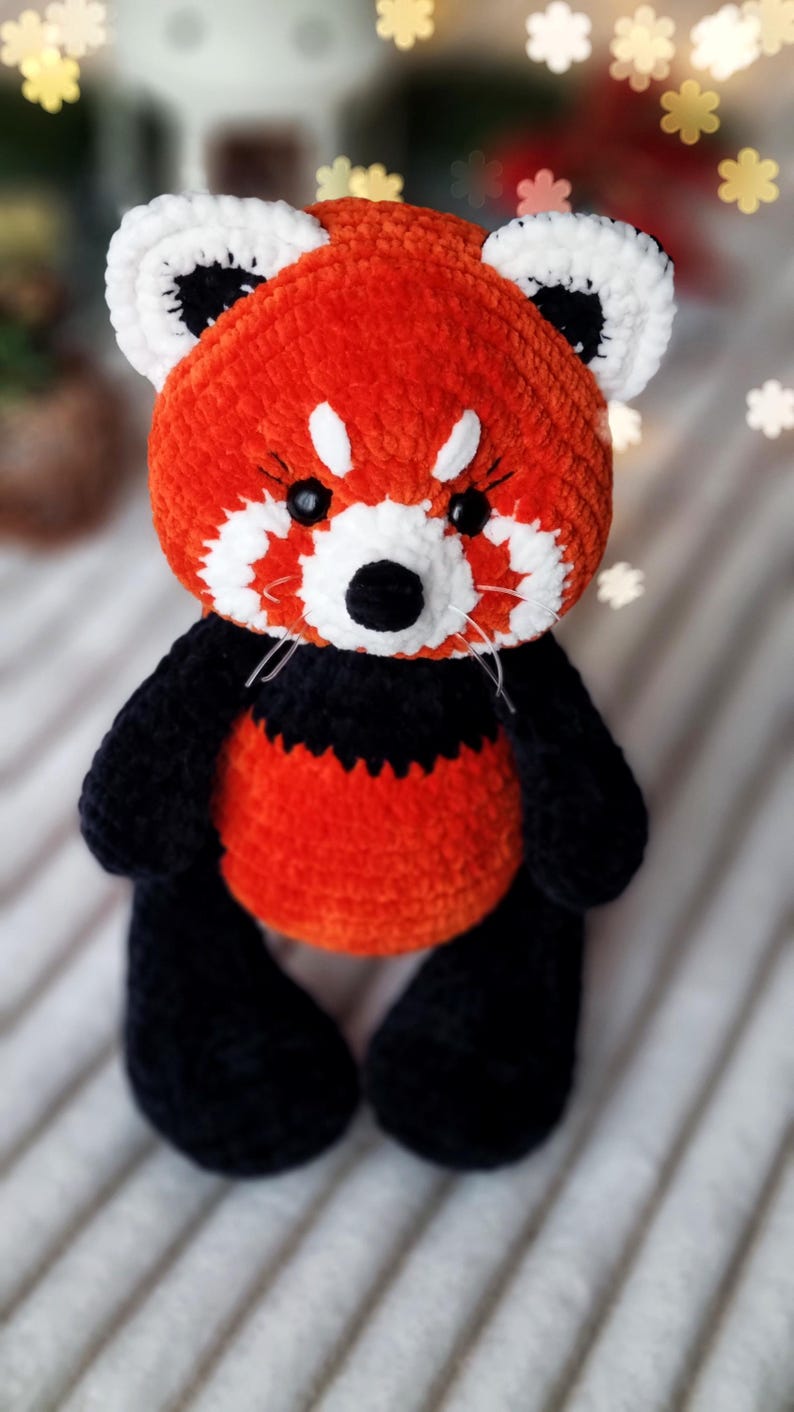 May include: A crocheted red panda plush toy with black and white details. The toy has a red body, black legs and ears, and white facial markings. The toy is sitting on a white fuzzy surface.