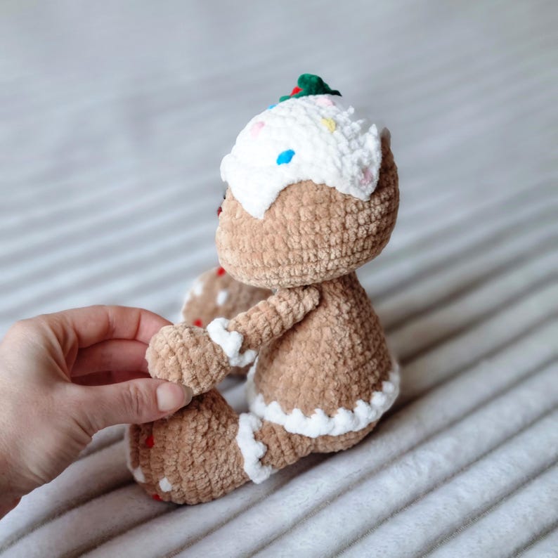 May include: A crocheted gingerbread man toy with brown fur, white icing, and colorful sprinkles. The toy is sitting on a white fluffy surface.