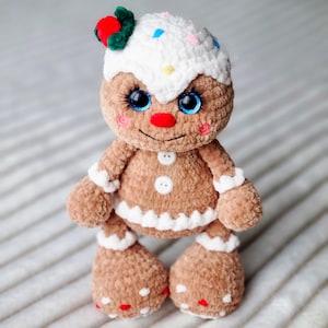 May include: A crocheted gingerbread man toy with white icing, red and white sprinkles, and blue eyes. The toy is wearing a red and green holly sprig on its head.