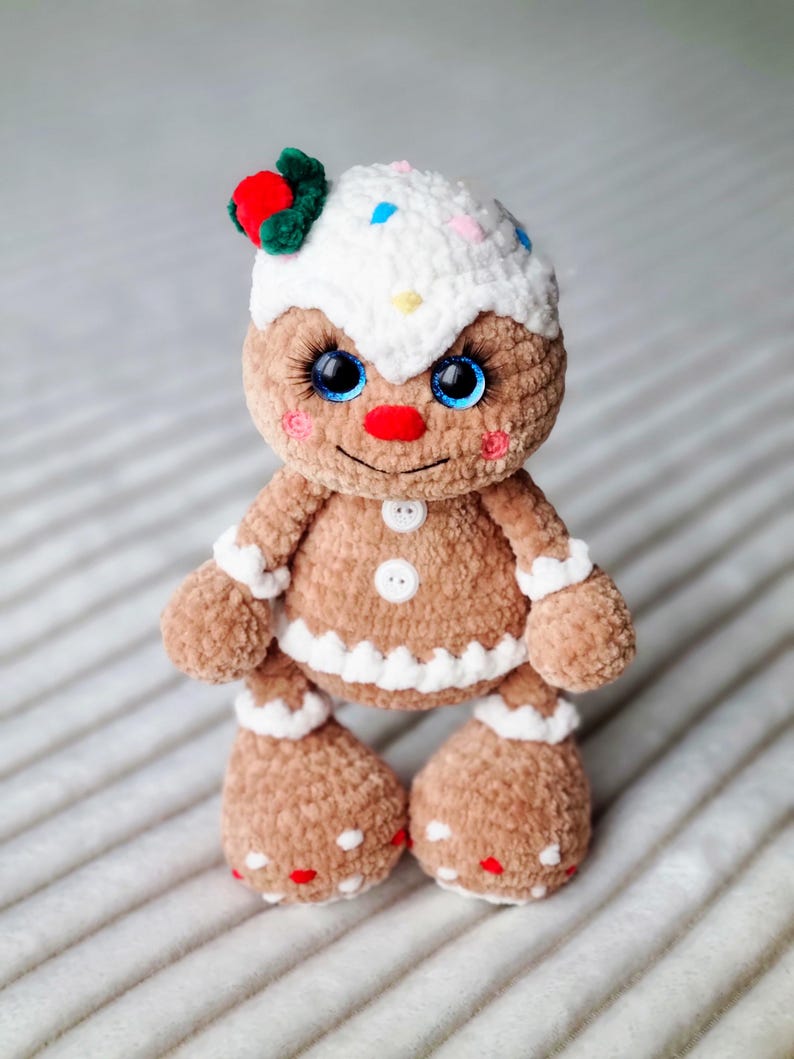 May include: A crocheted gingerbread man toy with white icing, red and white sprinkles, and blue eyes. The toy is wearing a red and green holly sprig on its head.