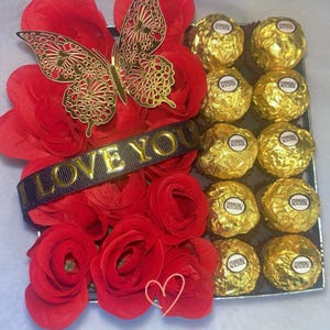 May include: A red rose arrangement with a gold butterfly and a black ribbon with the text "I LOVE YOU". The arrangement is next to a box of gold chocolate truffles.