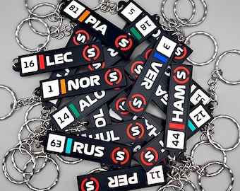 Puede incluir: Una colección de llaveros rectangulares negros con texto y números blancos que representan los nombres y números de pilotos de Fórmula 1, incluyendo PIA 81, LEC 16, NOR 1, VER 3, HAM 44 y RUS 63, cada uno con una anilla de metal plateado.