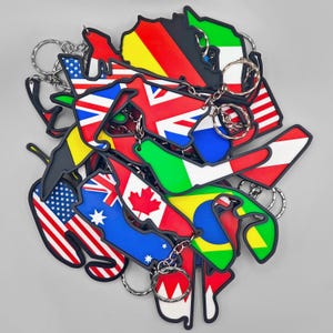 May include: A collection of Formula 1 racetrack keychains, each shaped like a specific circuit and filled with the colors of a national flag. The keychains feature metal rings and include designs representing countries like the USA, Canada, and Australia.