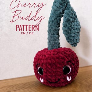 May include: A handmade, plush cherry toy with a smiling face, large, sparkly eyes, and a green stem. The text "Cherry Buddy PATTERN EN / DE" is written in a cursive font above the toy.