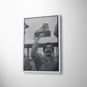 May include: Monochrome framed print showing a man holding a Nike shoe box. The box displays the Nike logo. The man, wearing a jumpsuit, has a moustache. The image is a vintage photograph.