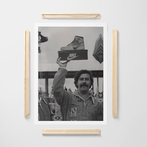 May include: Monochrome framed print showing a man holding a Nike shoe box with a high-top trainer. The man wears a jumpsuit and has a moustache. A crowd of people is visible in the background.