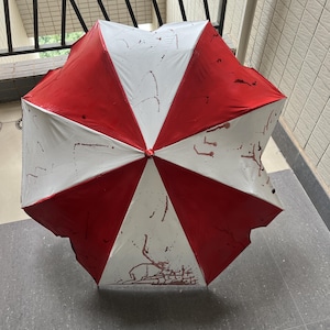 May include: A red and white umbrella with a segmented design. The umbrella has red panels alternating with white panels. There are dark red splatters on the white panels. The umbrella is open and resting on a gray surface.