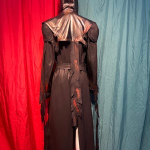 May include: A black hooded cloak with a leather-like appearance, featuring red accents and a belt. The cloak has a long, flowing design with jagged edges and is displayed against a red and teal backdrop.