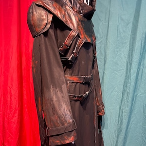 May include: Black costume with a white mask and a hood. The costume has a leather-like shoulder piece and accents with red paint splatters. The costume has buckles and straps. The costume is for Halloween or cosplay.