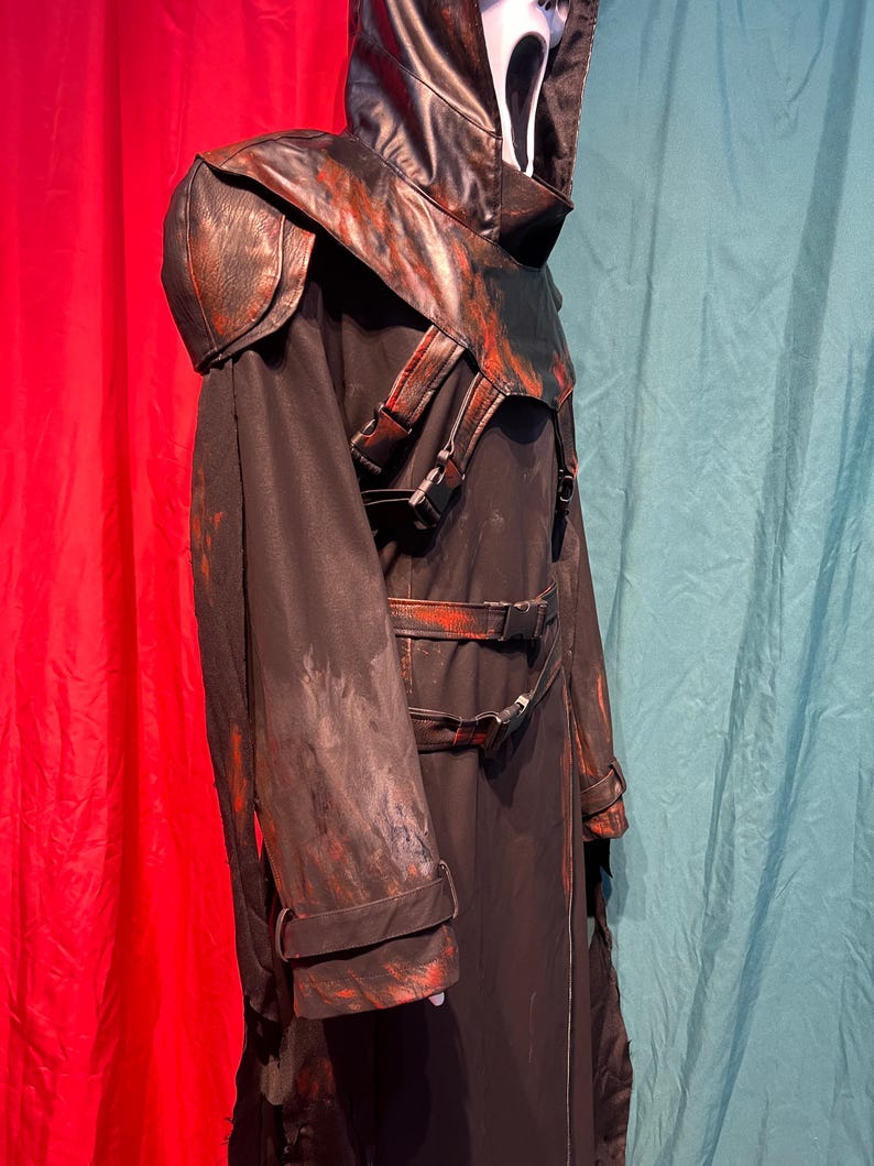 May include: Black costume with a white mask and a hood. The costume has a leather-like shoulder piece and accents with red paint splatters. The costume has buckles and straps. The costume is for Halloween or cosplay.