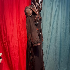 May include: A black and brown Scream costume with a white mask. The costume features a long, hooded robe with leather accents on the shoulders and hood. The robe has a tattered hemline and is secured with buckles.