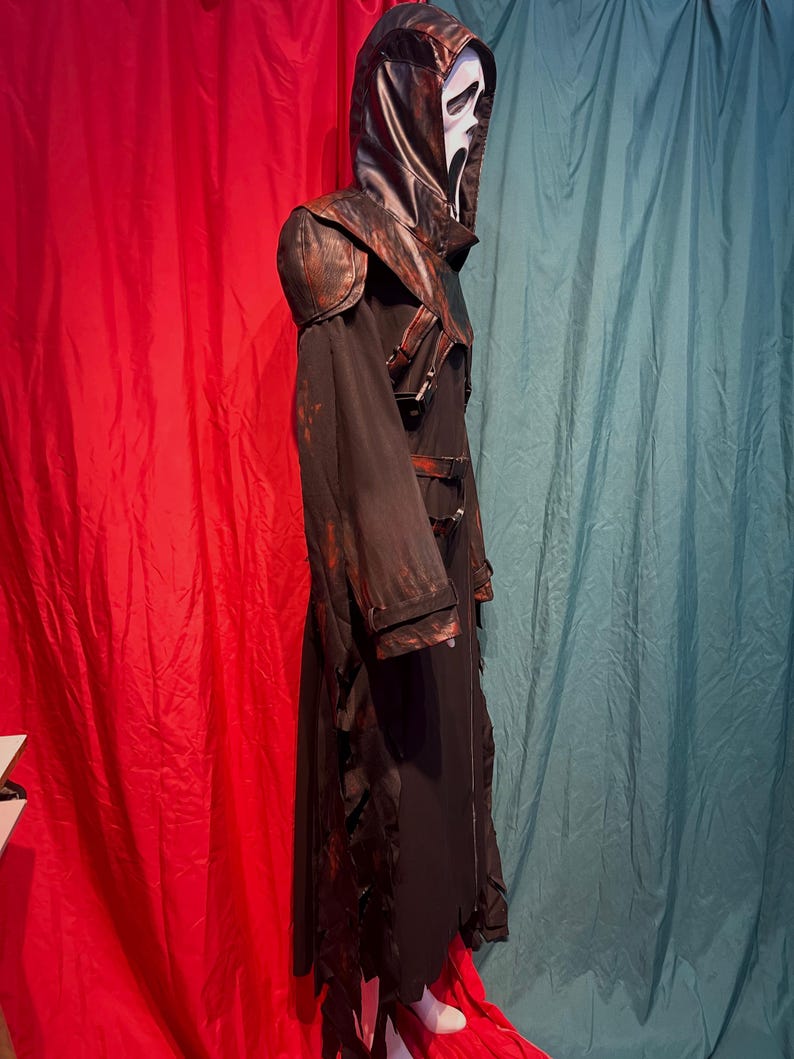 May include: A black and brown Scream costume with a white mask. The costume features a long, hooded robe with leather accents on the shoulders and hood. The robe has a tattered hemline and is secured with buckles.