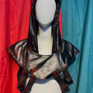 May include: A black faux leather hood and shoulder piece with red accents, displayed on a mannequin. The hood has a pointed top and the shoulder piece has straps with buckles. The background is red and teal.
